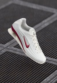 Bijela atletska cipela s teksturiranim gornjim dijelom i maroon akcentima, koja se ističe istaknutim swoosh logotipom, vezicama i elegantnim, modernim dizajnom.