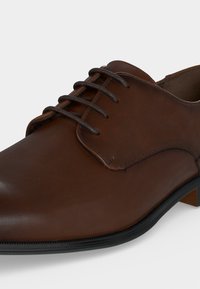 Chaussure formelle en cuir marron avec semelle noire et lacets marron foncé, gros plan montrant le dessus et les détails de la face avant.