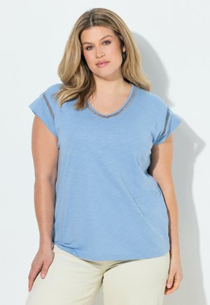 Kvinde med skulderlangt blondt hår iført en lyseblå kortærmet top med udskæringsdetaljer og cremefarvede bukser, stående foran en hvid baggrund.