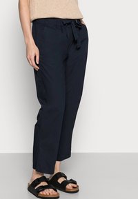 Kvinna i marinblå cropped byxor med midjeband, beige stickad tröja och svarta sandaletter med spännen stående mot en neutral bakgrund.