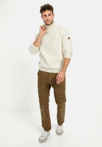 Cremfarbener Rippen-Turtleneck-Pullover, braune Cordhosen und weiße Sneaker. Auffällige Details: strukturierter Stoff, lockere Passform und kontrastierende Farben.
