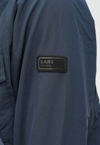 Granatowa kurtka o gładkiej fakturze, z czarną skórzaną naszywką na rękawie z napisem "BARBOUR INTERNATIONAL".