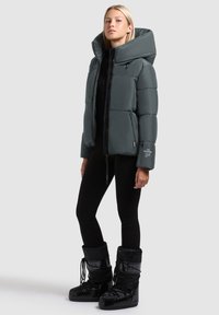 Kvinde iført en grå puffjakke med en overdimensioneret hætte, sorte leggings og sorte snesko med snørebånd, der står opad et ensfarvet baggrund.