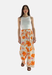 Witte tanktop gecombineerd met losse, bloemenpatroon broek in oranje en crème, met een elastische tailleband. Bruine sandalen maken de look af.