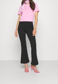 Kvinna iklädd en rosa kortärmad topp, svarta utsvängda byxor, rosa klacksandaler och en rosa- och vitmönstrad scarf på en vit bakgrund.