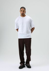 Wit katoenen t-shirt met korte mouwen, gecombineerd met een bruine broek en zwarte schoenen. De stof is glad; de schoenen hebben een veter met een contrasterende kleur.