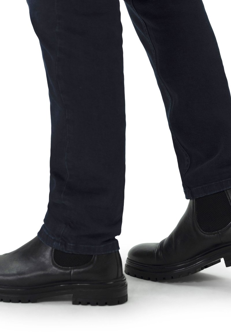 Zwarte leren enkellaarzen met textuuraccenten en meshpanelen, gecombineerd met donkerblauwe tapered broeken, zichtbaar vanuit een zijprofiel.