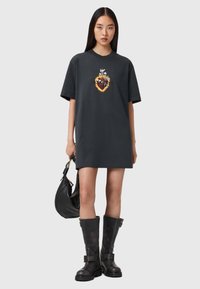 Czarna, oversize sukienka t-shirt z kolorową grafiką przedstawiającą serce i twarz, zestawiona z czarnymi, wysokimi na kolana butami oraz czarną torebką.
