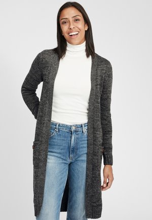 Cardigan grigio a maglia con frontale aperto e maniche lunghe, dotato di tasche laterali e tessuto testurizzato, indossato sopra un dolcevita bianco e jeans blu.