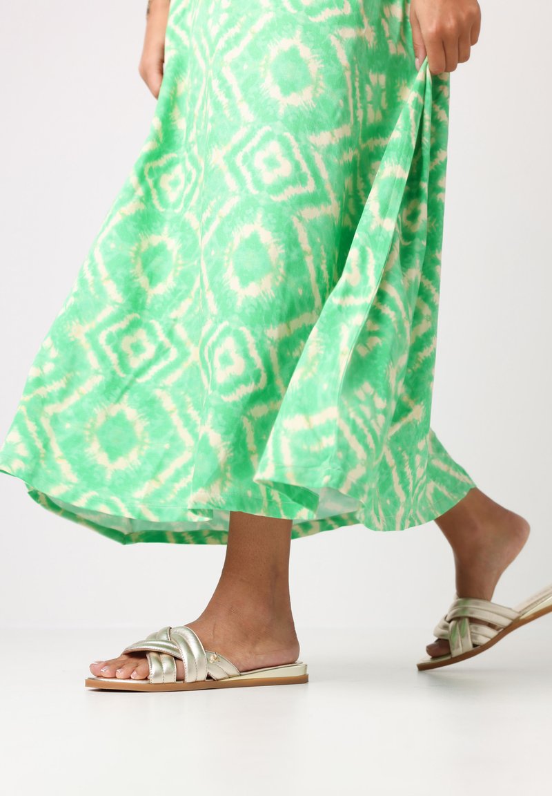 Robe maxi verte à motif avec un effet tie-dye et une main tenant l'ourlet ; associée à des sandales glissantes métalliques en tressé.