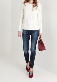 Suéter blanco de punto trenzado, jeans ajustados de color azul oscuro, tacones rojos de punta y un bolso burdeos. Texturas suaves y patrones superpuestos.