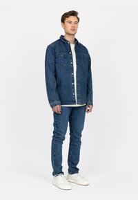 Denimjack met twee zakken aan de voorkant, knoopsluiting en een klassieke kraag, gecombineerd met slim-fit denimjeans en witte sneakers.