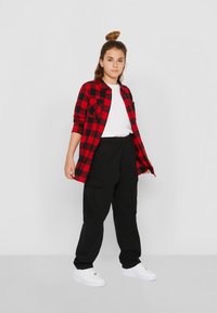 Chemise en flanelle à carreaux rouges et noirs, t-shirt blanc, pantalon cargo noirs et baskets blanches. Le modèle se tient les mains sur les hanches sur un fond gris.
