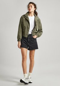 Chaqueta bomber verde oliva con puños elásticos, camiseta blanca, pantalones cortos de mezclilla negra de cintura alta y zapatillas blancas con detalles grises.