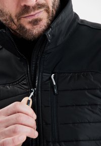 Sort jakke med en quiltet front, glatte stofærmer og kontrasterende lynlåse. Nærbillede viser en hånd, der griber fat om lynlåsens træk.