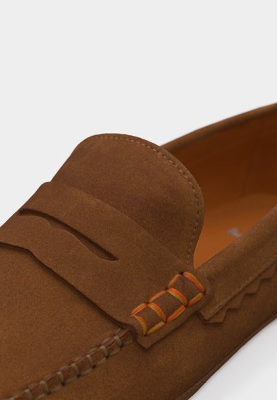 Mocassin en daim marron avec des détails en fil orange et jaune sur le côté, une couture de style mocassin le long du bord, et une lanière à rabat sur le dessus.