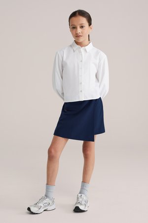 Pige står med hænderne bag ryggen iført hvid skjorte med knapper, marineblå nederdel, grå sokker og hvide sneakers på ensfarvet baggrund.