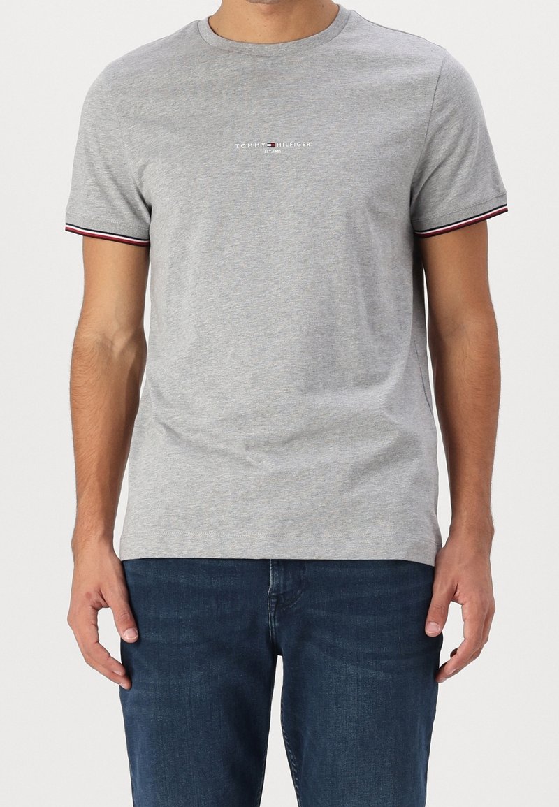 Harmaa puuvillainen T-paita, jossa on pyöreä kaula-aukko ja lyhyet hihat. Raitakoristeet punaisena ja sinisenä hihoissa. Keskellä logo-teksti vastakkaisissa väreissä.