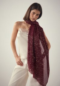 Bordeauxroter Schal mit goldenen Sprenkeln, aus leichtem Stoff, elegant um den Hals drapiert, mit einer durchscheinenden Struktur und weichem Finish.
