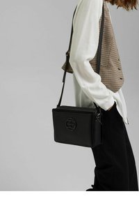 Una persona che indossa un gilet a quadri e una camicia bianca porta una borsa a tracolla rettangolare nera con un logo circolare davanti.