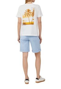 T-shirt blanche avec un graphique orange représentant des palmiers et du texte, associée à un short roulé bleu clair et des baskets blanches avec des accents noirs.