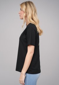 Schwarzes Baumwoll-T-Shirt mit kurzen Ärmeln, rundem Halsausschnitt und lässiger Passform. Mit einem kleinen grünen Grafikdesign auf der linken Rücken-Seite.