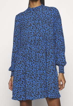 Frau trägt ein langärmliges, blaues Kleid mit Leopardenmuster, geraffter Taille und smockbündchen, steht vor einem weißen Hintergrund.