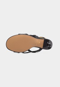 Μαύρο σανδάλι με λεπτά λουράκια και ψηλό τακούνι, με μπεζ σόλα, χρυσό λογότυπο και στρογγυλεμένη άκρη τακουνιού, εμφανιζόμενο από κάτω.