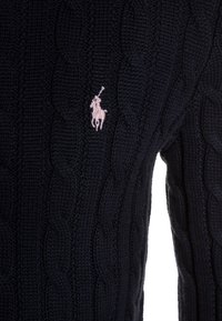 Marinblå kabelstickad tröja med en diskret rosa broderad polo-logotyp. Texturen är ribbad med intrikata kabelstickningsdetaljer.