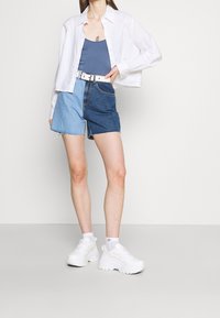 Przycięta biała koszula z guzikami, niebieska koszulka na ramiączkach, dżinsowe szorty z jasnymi i ciemnymi odcieniami, białe trampki z grubą podeszwą i białe skarpetki.