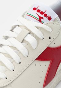 White leather sneaker with red accents, featuring flat white laces, a textured surface, and Diadora logo on the tongue.