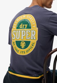 Donkergrijze t-shirt met een grote kleurrijke cirkelvormige grafiek op de achterkant met de tekst "SUPER," "dry," en "PREMIUM QUALITY" in verschillende kleuren.