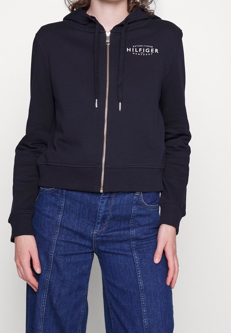 Femme portant un sweat à capuche noir zippé avec le logo « Hilfiger » sur la poitrine et un jean bleu taille haute, debout devant un fond uni.