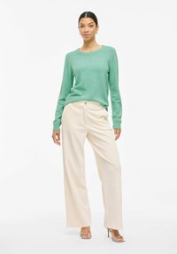 Una modelo se presenta con confianza, usando un suéter verde menta y pantalones de pierna ancha en color crema, complementados con sandalias de tacón plateadas.