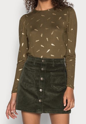 Femme portant un haut à manches longues vert olive avec imprimé plume dorée et une mini-jupe en velours côtelé vert foncé avec boutons à l'avant et poches devant.