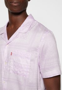 Gros plan d'une personne portant une chemise à manches courtes violet clair boutonnée avec un motif paisley blanc subtil et une poche poitrine avec une petite étiquette rouge.