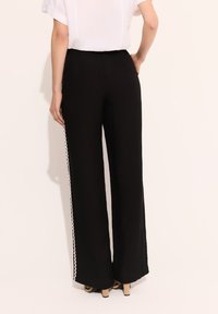 Czarne spodnie w szerokim kroju, o gładkiej teksturze, z białym falistym wykończeniem wzdłuż boku. Pas jest elastyczny, co zapewnia luźny krój.