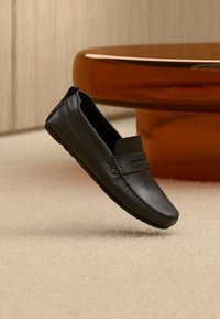 Μαύρο δερμάτινο παπούτσι τύπου loafer με ραφές, μικρή ετικέτα με λογότυπο, τοποθετημένο σε μπεζ χαλί κοντά σε στρογγυλό καφέ τραπεζάκι.