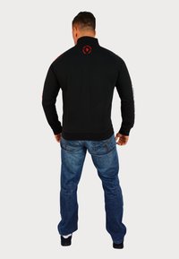 Felpa nera con zip e collo alto, con dettagli rossi e logo sul retro. Indossata con jeans blu e scarpe nere.