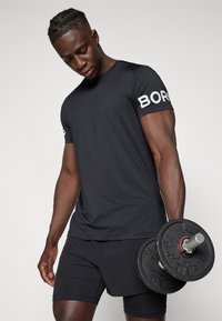 Schwarzes sportliches T-Shirt mit "BORG" in Weiß auf dem Ärmel, kombiniert mit dunklen Shorts. Die Person hält eine schwarze Hantel.