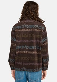 Persona con lunghi capelli ricci che indossa una giacca in pile marrone con motivi astratti blu, verdi e viola e jeans blu, girata di spalle.