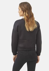 Sudadera negra con un panel de malla texturizada en la cintura, con un ajuste relajado y puños anchos. El cabello es ondulado y largo, peinado hacia abajo.