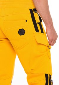 Gelbe Baumwoll-Cargohosen mit schwarzen Seitenstreifen, mehreren Taschen und hexagonalem Logo-Patch auf der hinteren Tasche. Funktionale Designdetails sichtbar.