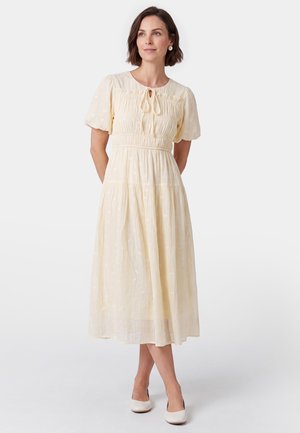 Vrouw in een crèmekleurige midi-jurk met pofmouwen, een strikkoord in de hals en witte schoenen, staand tegen een effen achtergrond.