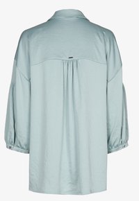 Blouse bleu clair à manches longues, coupe ample, pli plat dans le dos et poignets boutonnés, vue de dos.