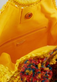 Wnętrze żółtej plecionej torby z gładką podszewką, zawierające kieszonkę na zamek błyskawiczny oraz ozdobny pompon w żywych kolorach po boku.