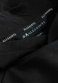 Tessuto nero che mostra un primo piano di un'etichetta con le parole "ALLSAINTS" e "SMALLSAINTS" in carattere bianco a contrasto contro il materiale testurizzato.