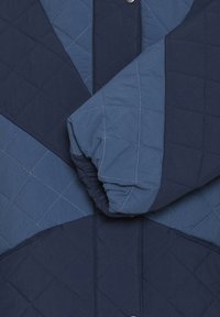 Chaqueta acolchada en diferentes tonos de azul, con motivos en diamante y un puño recogido, que presenta un detalle de botón a presión visible.