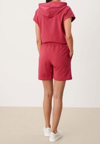 Czerwona krótka bluza z kapturem, z elastycznym pasem i kieszeniami, wykonana z miękkiego materiału; zestawiona z białymi trampkami.