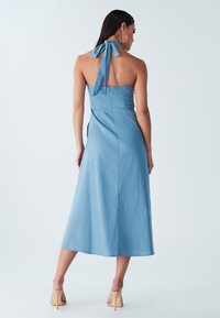 Naine seisab seljaga vaataja poole, kandes helehalli sinist ilma varrukateta midi pikkusega kleiti ja beeže kõrge kontsaga sandaale, taust on lihtne.